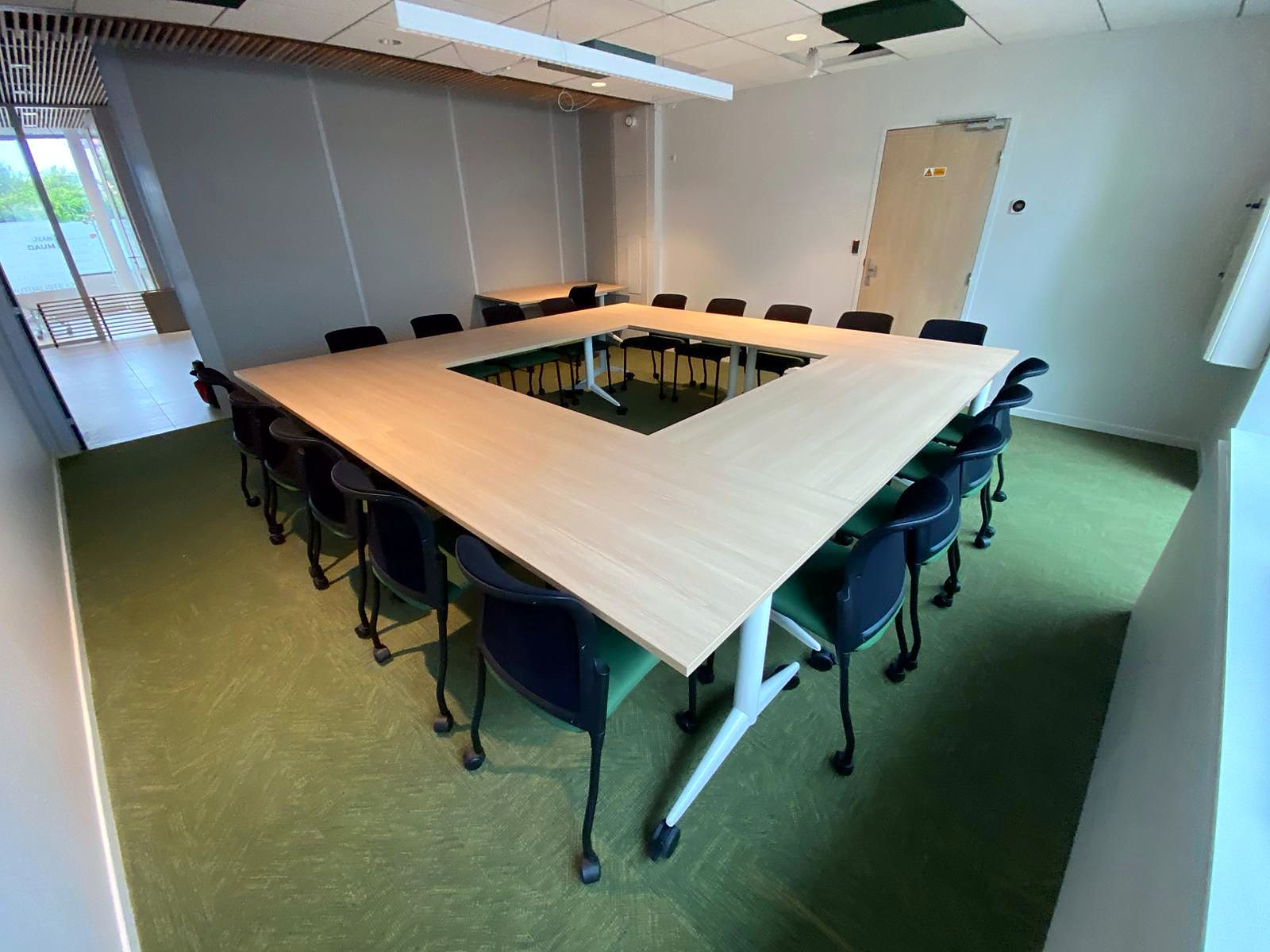 Salle de formation : tables assemblées en carré ouvert, sièges noirs empilables, moquette verte