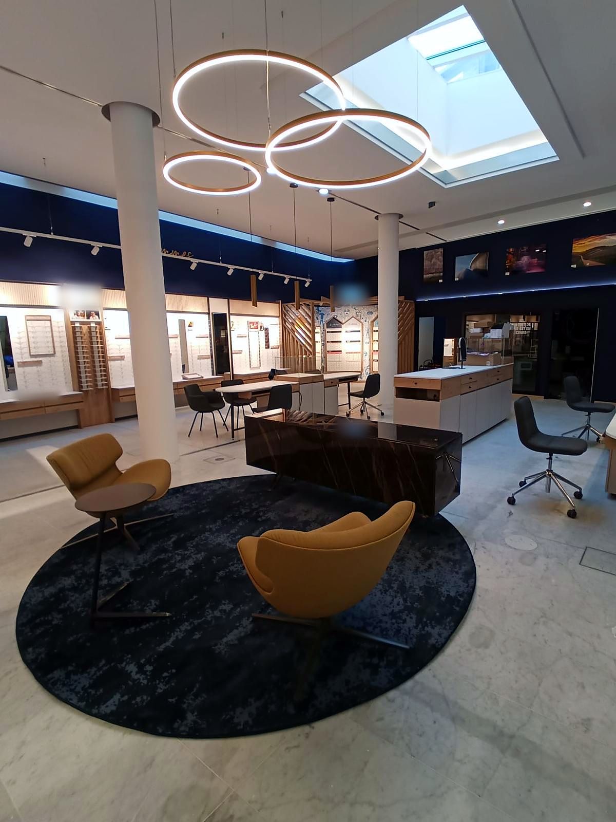 Espace d’accueil : fauteuils cognac sur tapis rond, suspensions annulaires et verrière zénithale